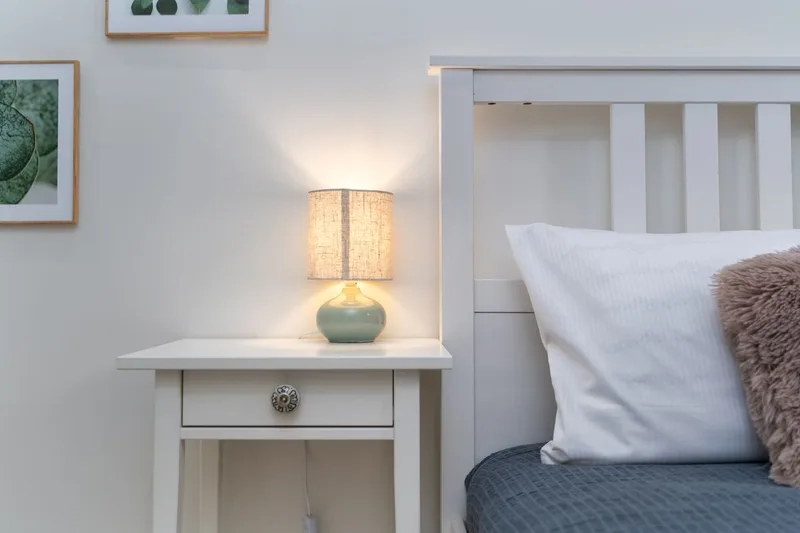 A softly lit bedside lamp on a white nightstand beside a neatly made bed, conveying privacy and calm consideration